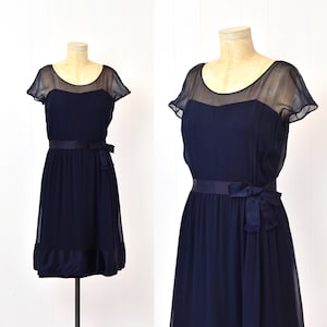 May include: Navy blue dress with a sheer overlay on the bodice and sleeves. The dress features a fitted waist with a matching sash tied in a bow. The skirt is a flowing style, and the dress is displayed on a mannequin.