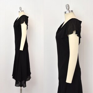 May include: A black vintage dress with a V-neck and short, flutter sleeves. The dress has a fitted bodice and a flared skirt with a layered hem. The dress is displayed on a mannequin.