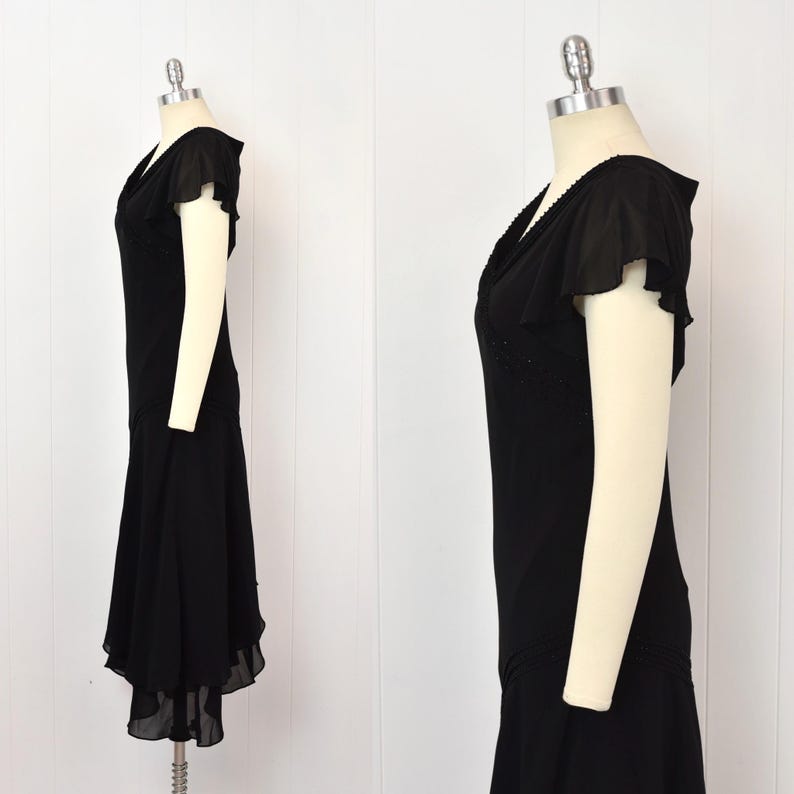 May include: A black vintage dress with a V-neck and short, flutter sleeves. The dress has a fitted bodice and a flared skirt with a layered hem. The dress is displayed on a mannequin.