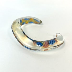 May include: A clear glass bangle bracelet with a colorful floral design inside. The bangle is curved and has a smooth, polished finish.