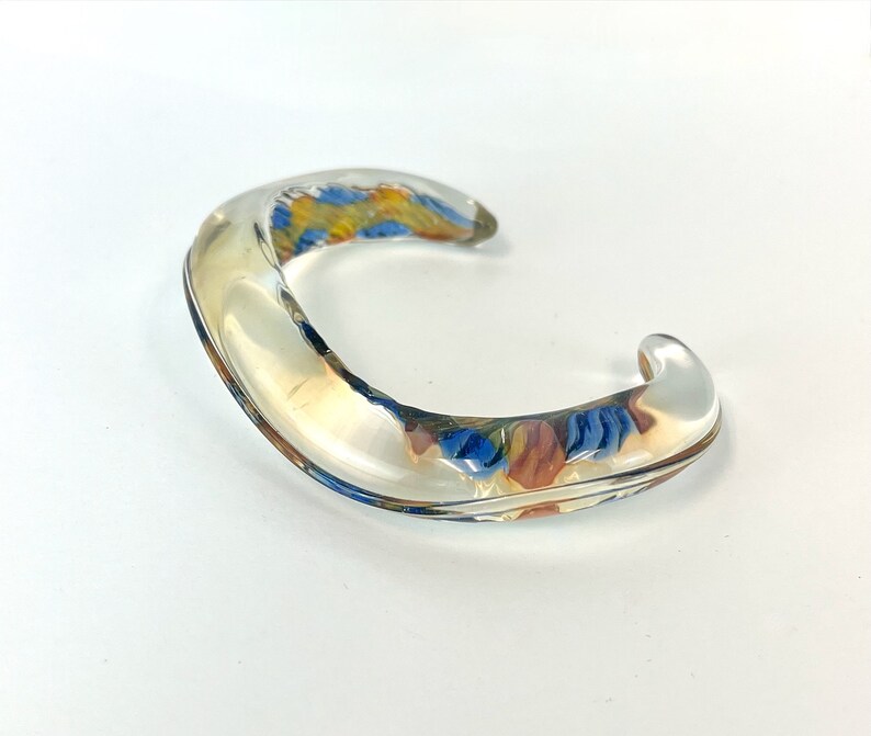 May include: A clear glass bangle bracelet with a colorful floral design inside. The bangle is curved and has a smooth, polished finish.