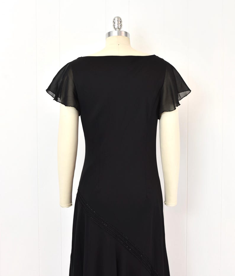 May include: A black dress with a square neckline and short, sheer flutter sleeves. The dress has a fitted bodice and a flared skirt. A diagonal line of small beads accents the front of the skirt. The dress is displayed on a mannequin.
