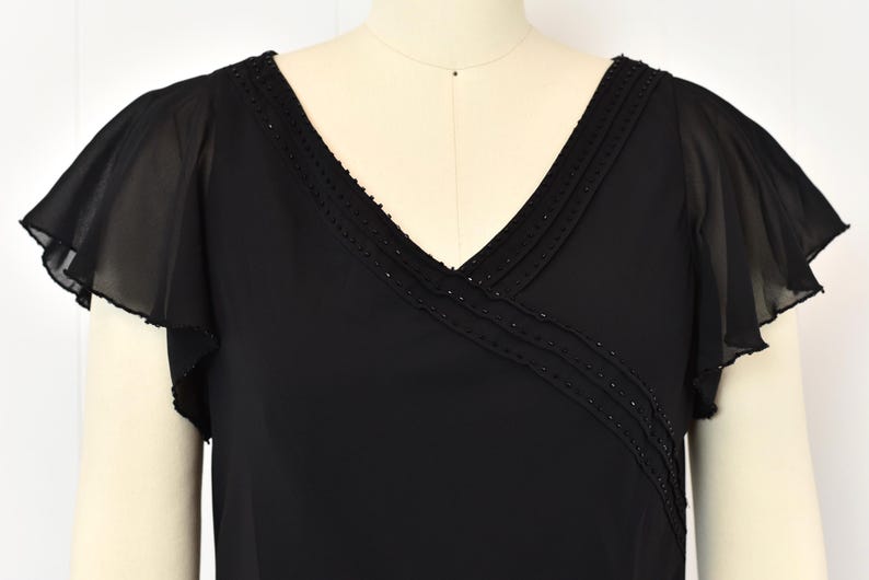 May include: Black V-neck top with flutter sleeves. The neckline and upper bodice feature a beaded trim. The sleeves are made of a sheer, flowing fabric. The top is displayed on a mannequin.