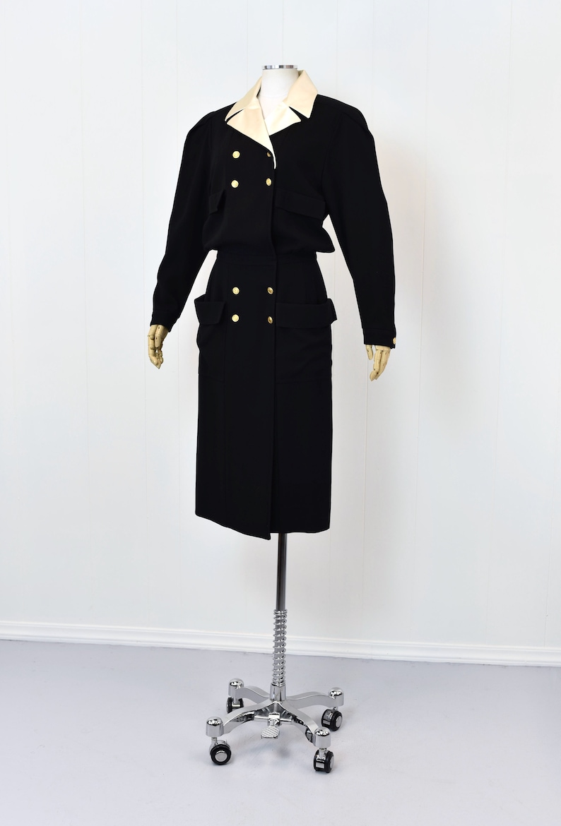 May include: A black dress with a white collar and gold buttons. The dress has a double-breasted closure and large pockets on the sides.