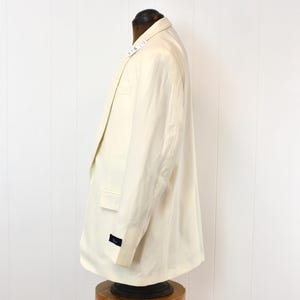 May include: Cream-colored blazer displayed on a mannequin. The blazer has a classic design with a notched lapel, a chest pocket, and two side pockets. The sleeves have a button detail. The fabric appears to be a lightweight material.