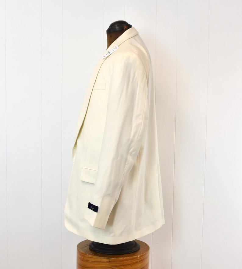May include: Cream-colored blazer displayed on a mannequin. The blazer has a classic design with a notched lapel, a chest pocket, and two side pockets. The sleeves have a button detail. The fabric appears to be a lightweight material.