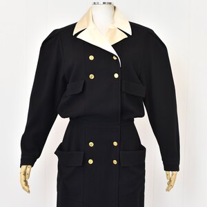 May include: A black double-breasted dress with a white collar and gold buttons. The dress has large pockets on the front and a long skirt.