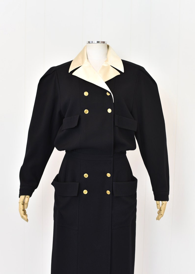 May include: A black double-breasted dress with a white collar and gold buttons. The dress has large pockets on the front and a long skirt.