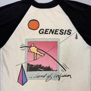 May include: Cream-colored raglan t-shirt with black sleeves. The shirt features the band name "GENESIS" above a graphic of a yellow figure, a pink and black cityscape, and the text "...land of confusion."
