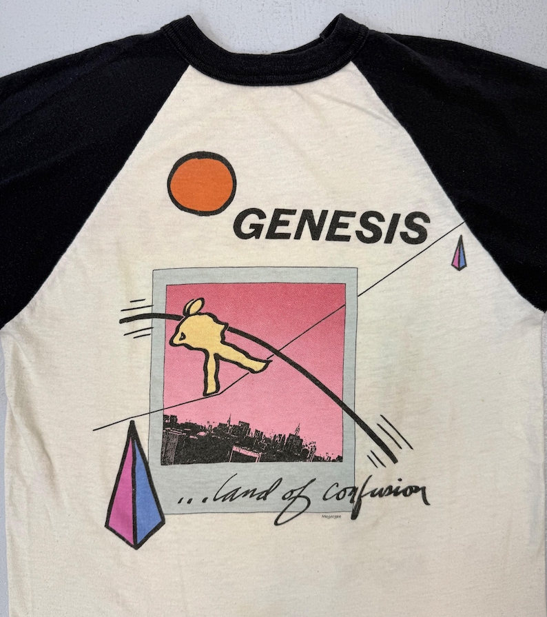 May include: Cream-colored raglan t-shirt with black sleeves. The shirt features the band name "GENESIS" above a graphic of a yellow figure, a pink and black cityscape, and the text "...land of confusion."
