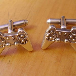 May include: A pair of silver-toned cufflinks shaped like video game controllers. The controllers have a detailed design with buttons and a directional pad.