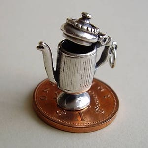 以下が含まれることがあります： 蓋付きのティーポットの形をした銀製のチャーム。チャームは銅色のコインの上に置かれています。