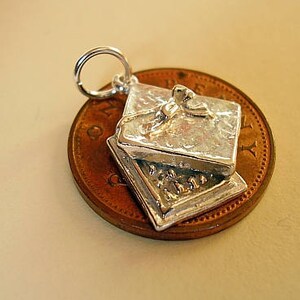 May include: Silver charm in the shape of a closed box with a bow on top. The charm is shown on a penny for size comparison.