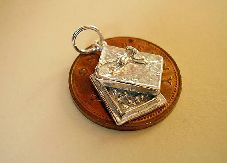 May include: Silver charm in the shape of a closed box with a bow on top. The charm is shown on a penny for size comparison.
