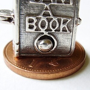 May include: Silver charm in the shape of a book with the word "BOOK" embossed on the front. The charm is sitting on a copper penny.