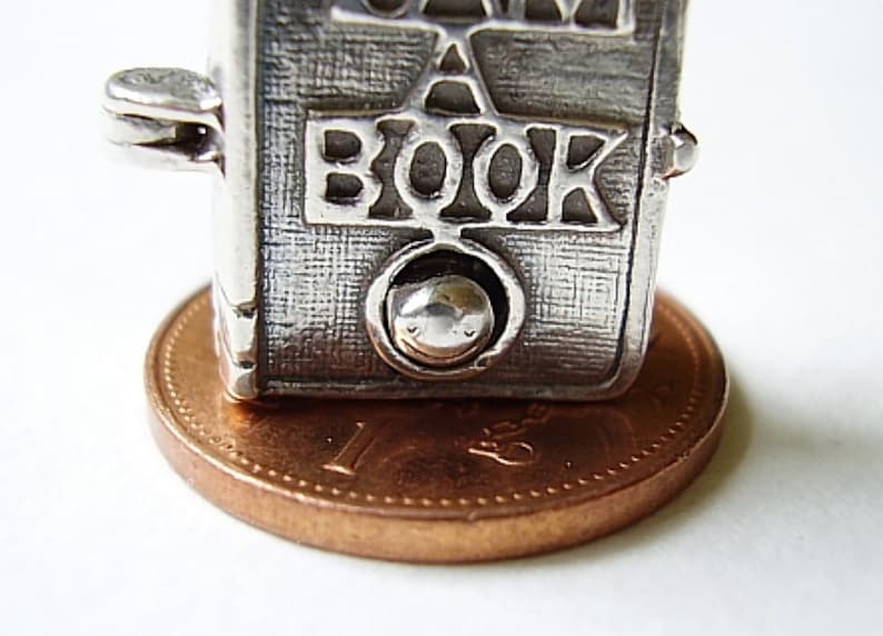 May include: Silver charm in the shape of a book with the word "BOOK" embossed on the front. The charm is sitting on a copper penny.