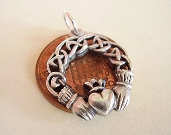 May include: A silver Claddagh pendant, with a heart, hands, and crown design, resting on a copper penny. The pendant has Celtic knot detailing. The penny provides a contrasting background to the silver pendant.