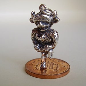 May include: Silver charm of a small, detailed, standing, mythical creature with horns, a furry body, and a human-like face. The charm is on top of a British penny.