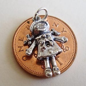 May include: Silver charm of a rag doll with a smiling face, wearing a dress and holding a bow in her hand. The charm is sitting on a copper penny.