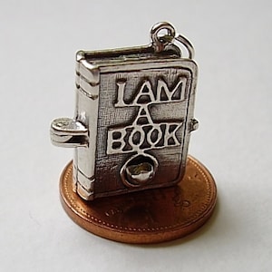 May include: Silver charm in the shape of a book with the words "I AM A BOOK" on the cover. The charm is sitting on a penny.