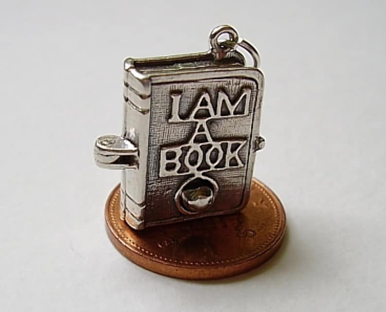 May include: Silver charm in the shape of a book with the words "I AM A BOOK" on the cover. The charm is sitting on a penny.