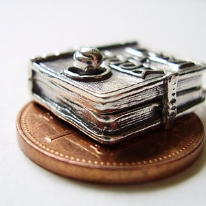May include: Silver charm in the shape of a closed book with a decorative knob on the top. The charm is sitting on a copper penny.