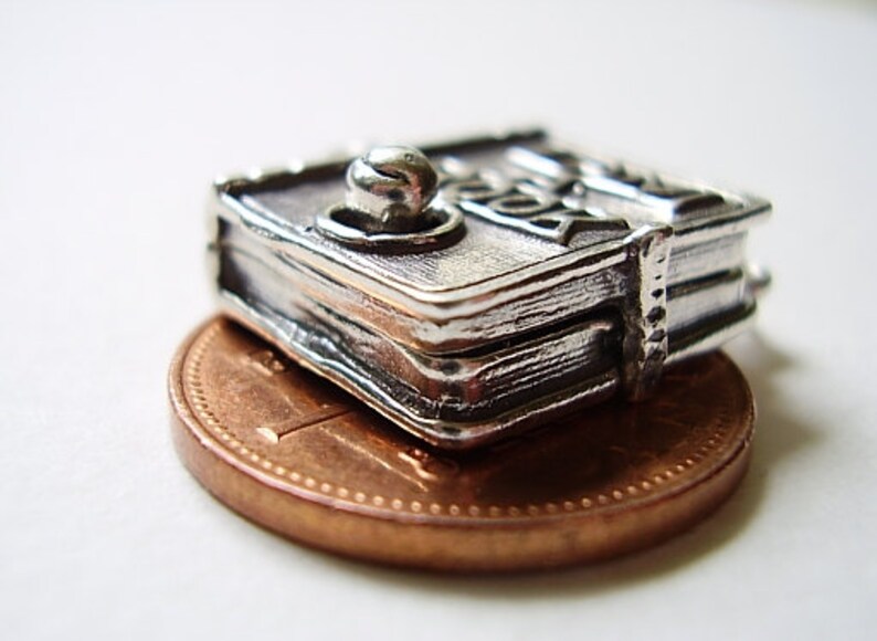 May include: Silver charm in the shape of a closed book with a decorative knob on the top. The charm is sitting on a copper penny.