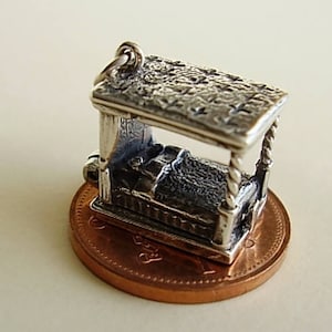 May include: A silver charm depicting a four-poster bed with intricate detailing. The charm is resting on a copper penny.