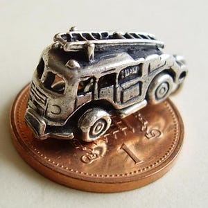 May include: A silver charm in the shape of a vintage fire engine with a ladder on top. The charm is sitting on a British one pence coin.