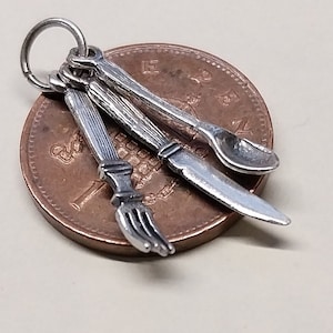 Op de afbeelding: Zilveren bedel met een mes, vork en lepel. De bedel is ongeveer zo groot als een Nederlandse 1-cent munt.