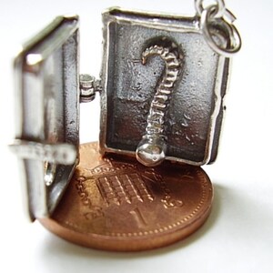 May include: Silver charm in the shape of a book that opens to reveal a silver skeleton inside. The charm is about the size of a US penny.