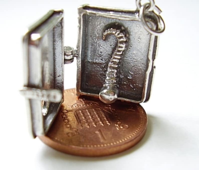 May include: Silver charm in the shape of a book that opens to reveal a silver skeleton inside. The charm is about the size of a US penny.