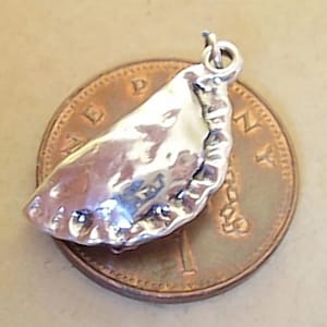 May include: A silver charm in the shape of a pasty, on a penny coin. The charm is detailed and has a textured surface.