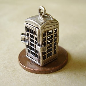 May include: A small, silver-colored charm in the shape of a British telephone box. The charm has a rectangular shape with a door and window details. The word "PHONE" is visible at the top. The charm is sitting on a penny.