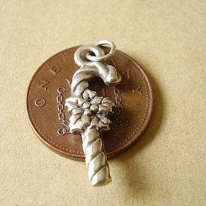 May include: Silver charm in the shape of a candy cane with a poinsettia flower design. The charm is sitting on a copper penny.