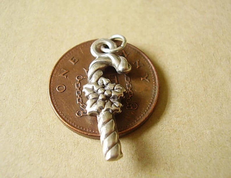 May include: Silver charm in the shape of a candy cane with a poinsettia flower design. The charm is sitting on a copper penny.