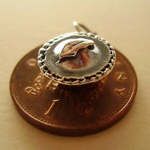 May include: Silver charm in the shape of a pie with a crust and a single cherry on top. The charm is sitting on a copper-colored coin.