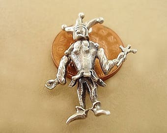 May include: A silver-colored charm in the shape of a jester, with a pointed hat and holding a small object in each hand. The jester charm is standing on a copper-colored coin.