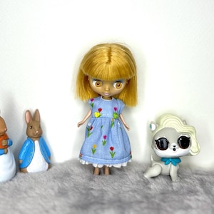 May include: A Blythe doll with blonde hair wearing a light blue dress embroidered with colorful flowers. Two rabbit figurines and a gray dog figurine with a blue bow are also in the image.