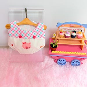 Pu&ograve; includere: Un indumento in miniatura su una gruccia di legno con un top a quadretti vichy rosa e verde acqua, due bottoni rossi e una parte inferiore bianca con un piccolo muso di maiale ricamato. Accanto, un carretto dei dolci giocattolo rosa con quattro cupcake.