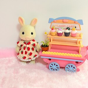 May include: A flocked rabbit figurine wearing a cream-colored dress with a red strawberry pattern, standing next to a pink and orange toy candy cart. The cart features two shelves holding four miniature cupcakes and a small potted daisy plant.