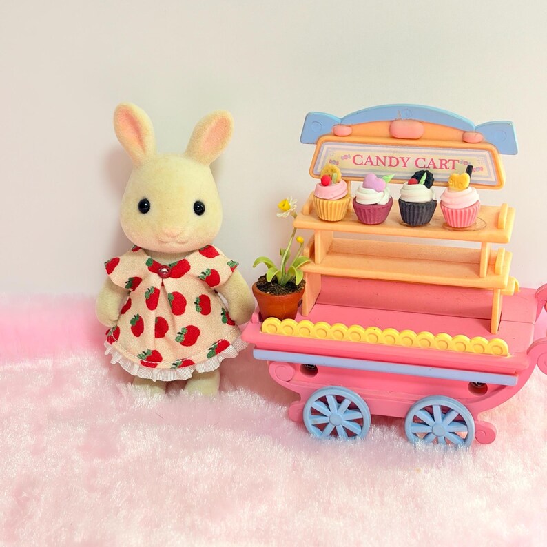 May include: A flocked rabbit figurine wearing a cream-colored dress with a red strawberry pattern, standing next to a pink and orange toy candy cart. The cart features two shelves holding four miniature cupcakes and a small potted daisy plant.