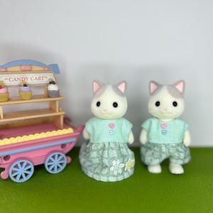 Puede incluir: Un carrito de caramelos de juguete rosa y azul, con un letrero que dice "CANDY CART" y tres cupcakes. Dos figuras de gato visten camisetas verdes y faldas estampadas. Las figuras de gato tienen pelaje gris y blanco.