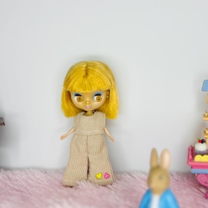 May include: A doll with yellow hair and a tan jumpsuit with heart-shaped patches. The doll stands on a pink surface, with a miniature candy cart and a toy rabbit in the background. A sewing machine is on the left.