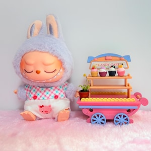 Pu&ograve; includere: Una bambola di peluche a forma di coniglio con il viso color pesca e una bavaglino bianco con un disegno di un maialino rosa, accanto a un carrello dei dolci giocattolo rosa con due ripiani, quattro ruote blu e quattro cupcake con la scritta CANDY CART.