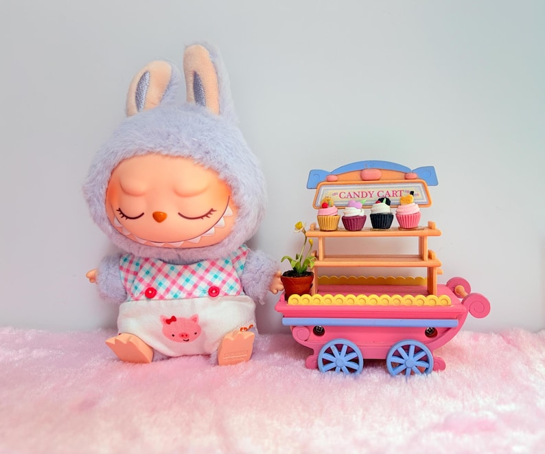 Pu&ograve; includere: Una bambola di peluche a forma di coniglio con il viso color pesca e una bavaglino bianco con un disegno di un maialino rosa, accanto a un carrello dei dolci giocattolo rosa con due ripiani, quattro ruote blu e quattro cupcake con la scritta CANDY CART.