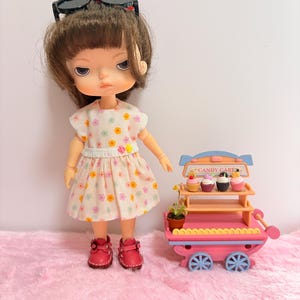 May include: A doll with brown hair wearing black sunglasses and a white dress with a colorful floral pattern, paired with red shoes. Beside the doll is a miniature pink and blue candy cart with four small cupcakes and a tiny potted plant.
