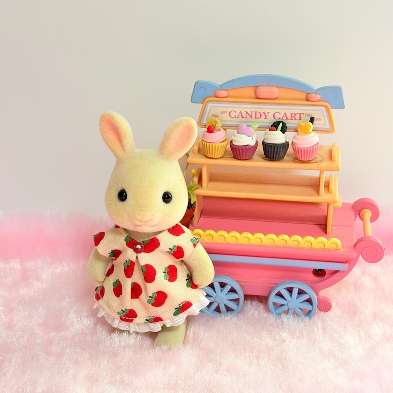 May include: A flocked cream-colored rabbit figurine wearing a beige dress with a red strawberry pattern, standing next to a pink and blue toy candy cart featuring two shelves stocked with four miniature colorful cupcakes and a sign reading CANDY CART.