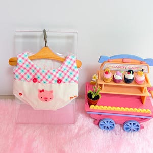 Pu&ograve; includere: Un indumento in miniatura con un top a quadri turchese e rosa, due bottoni rossi e una parte inferiore color crema con il disegno di una faccia di maialino rosa. Accanto c'&egrave; un carretto dei dolci in miniatura arancione e rosa con quattro cupcake.