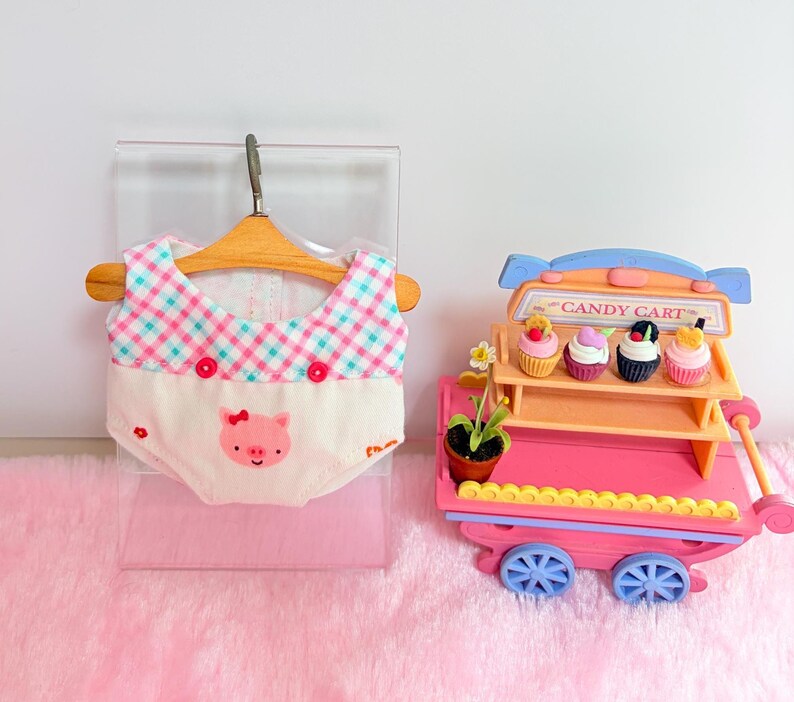 Pu&ograve; includere: Un indumento in miniatura con un top a quadri turchese e rosa, due bottoni rossi e una parte inferiore color crema con il disegno di una faccia di maialino rosa. Accanto c'&egrave; un carretto dei dolci in miniatura arancione e rosa con quattro cupcake.
